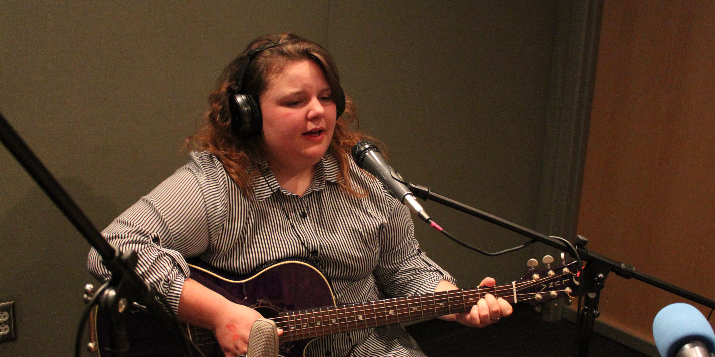 An adult wearing headphones playing acoustic guitar and singing into a microphone in a radio studio