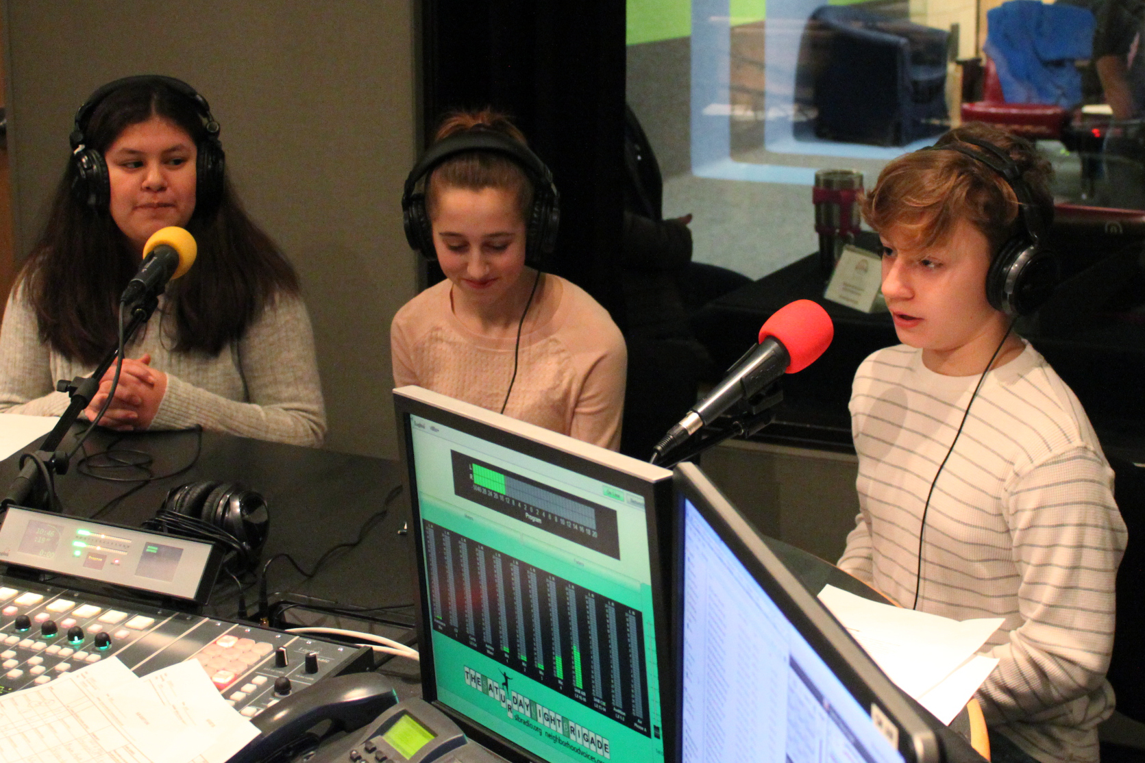 Three teens wearing headphones speaking into microphones being interviewed in a radio studio