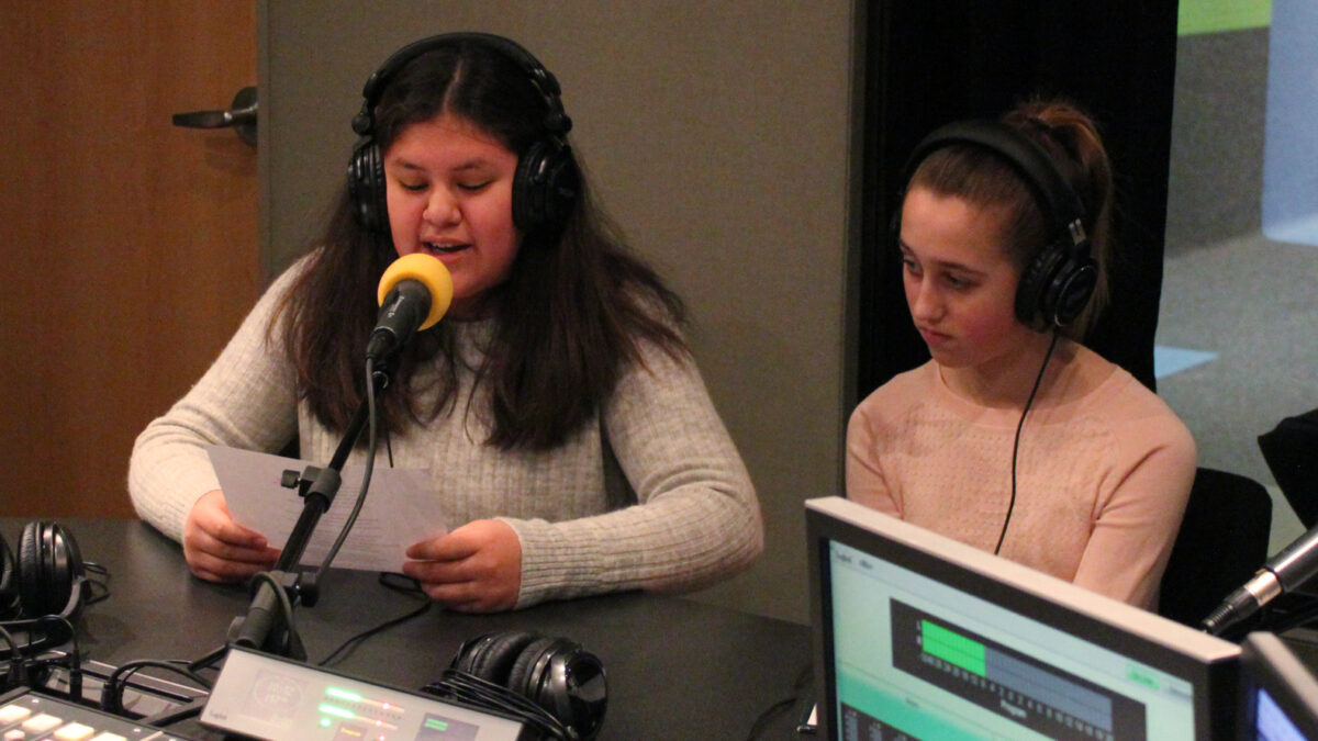 Two teens wearing headphones speaking into microphones in a radio studio
