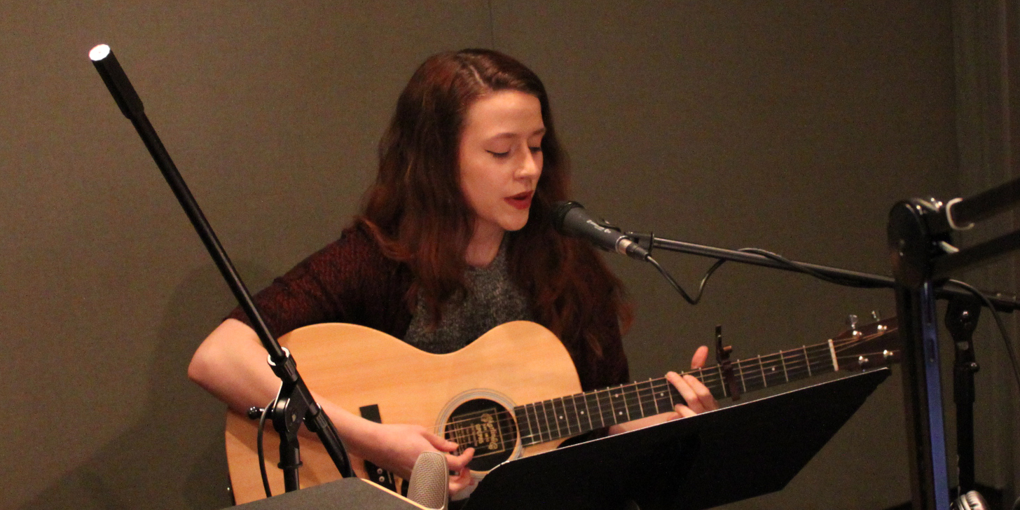 An adult playing an acoustic guitar and singing into a microphone in a radio studio