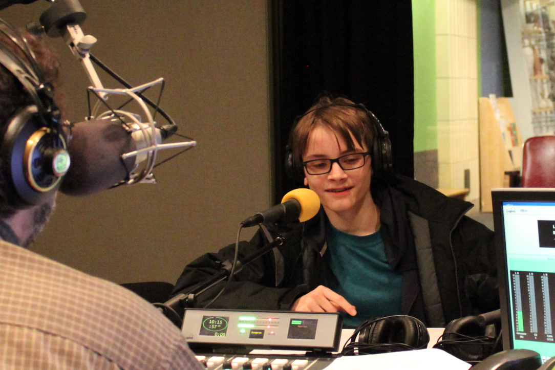 A teen wearing headphones speaking into a microphone being interviewed by an adult wearing headphones sitting behind a microphone in a radio studio