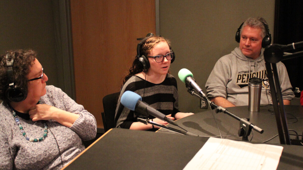 A teen wearing headphones speaking into a microphone as two adults wearing headphones listen in a radio studio