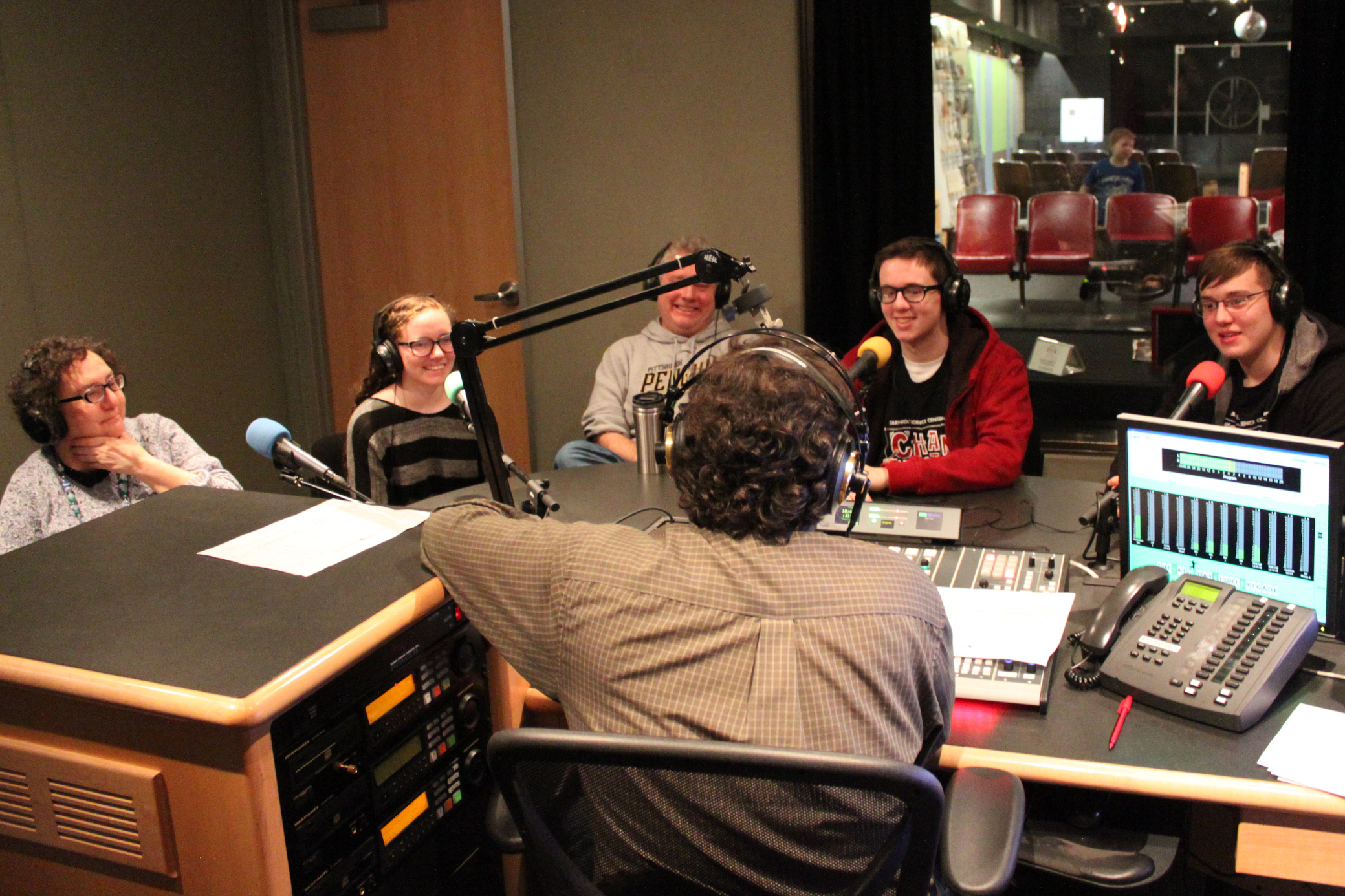 Three teens and two adults wearing headphones smiling behind microphones being interviewed by an adult wearing headphones sitting behind a mixing board in a radio studio