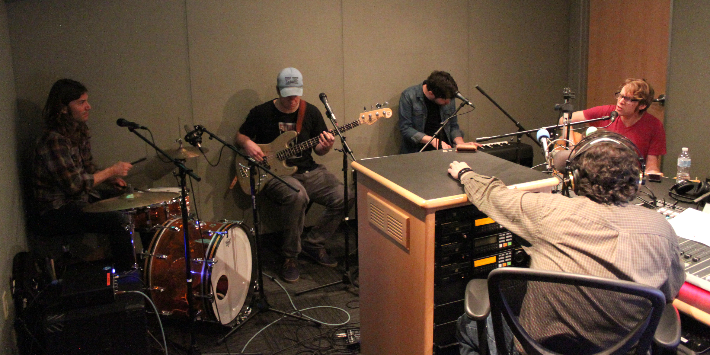 An adult playing a drum set, an adult playing electric bass, an adult playing a piano keyboard, and an adult singing into a microphone as another adult wearing headphones sitting behind a microphone and a mixing board listens in a radio studio