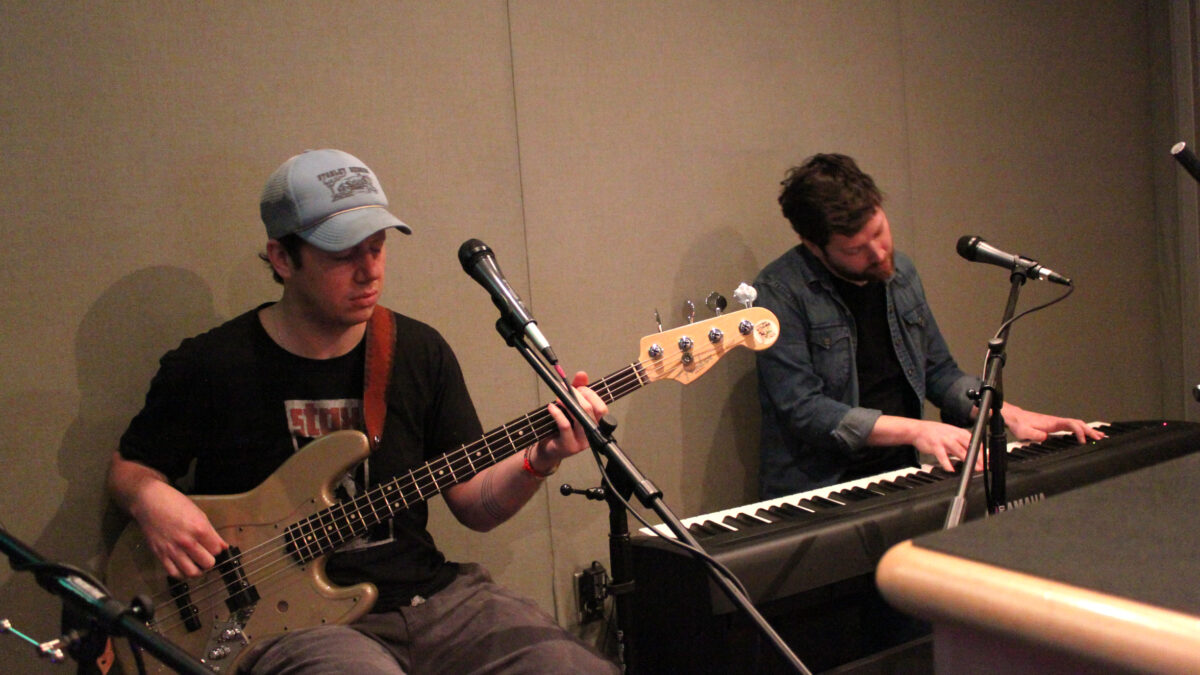 An adult playing electric bass and an adult playing a piano keyboard in a radio studio