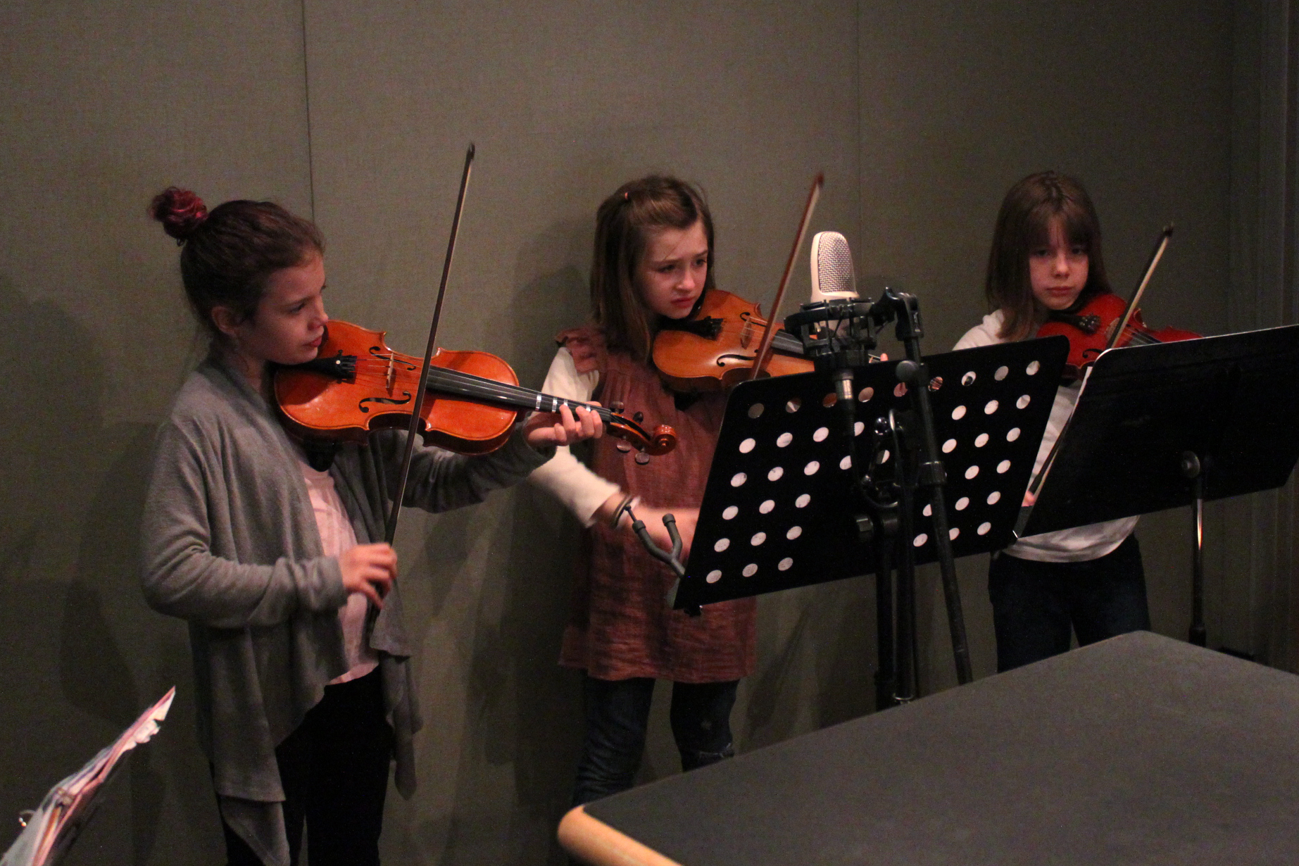 Three youth playing violin and looking at sheet music on music stands in a radio studio