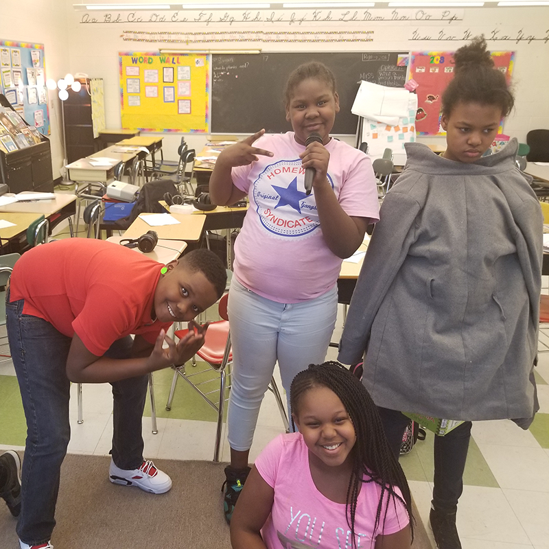 Four youth posing in a classroom, one holding a microphone