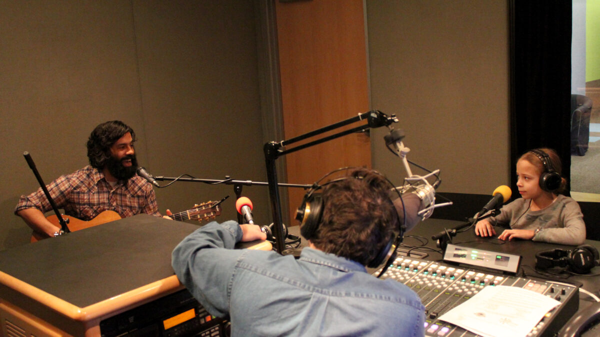 An adult playing acoustic guitar and singing into a microphone as a child wearing headphones and an adult wearing headphones and sitting behind a microphone and a mixing board listens in a radio studio