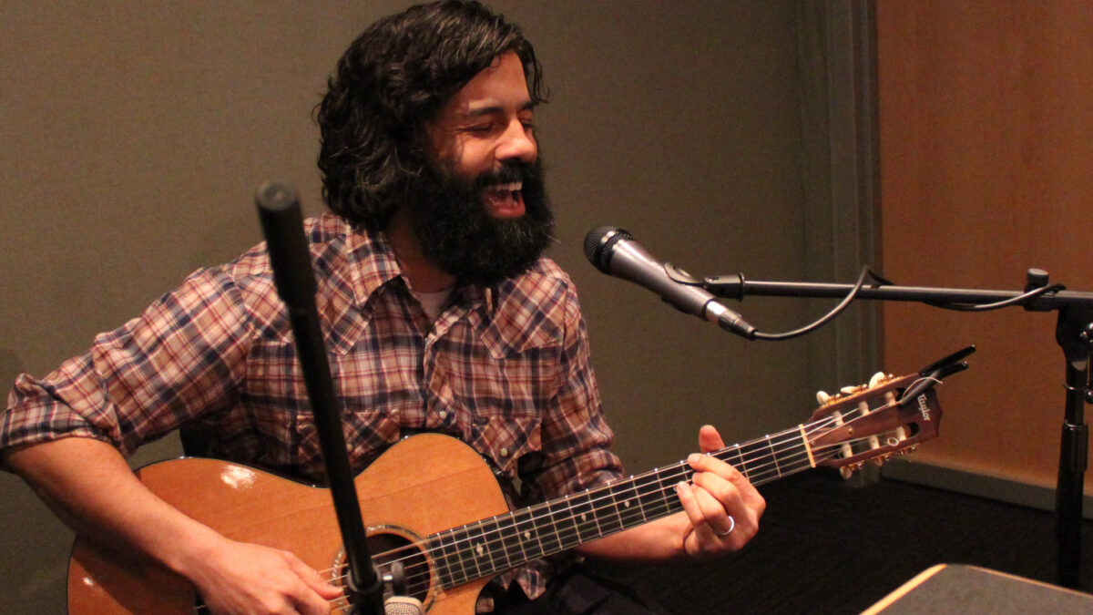 An adult playing acoustic guitar and singing into a microphone in a radio studio