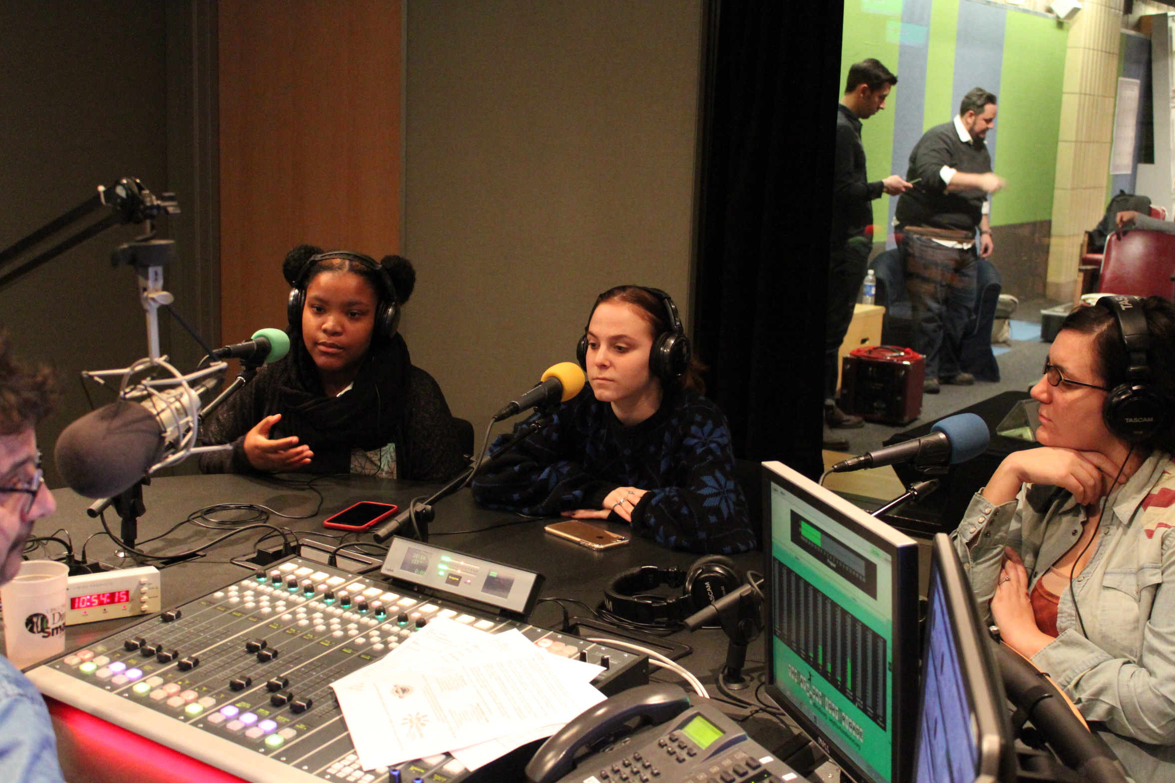 Three teens wearing headphones and speaking into microphones being interviewed by an adult sitting behind a microphone and a mixing board in a radio studio