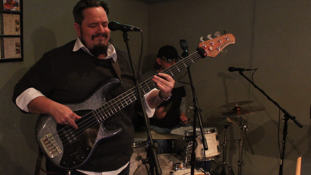 An adult playing electric bass and an adult playing a drum set in a radio studio