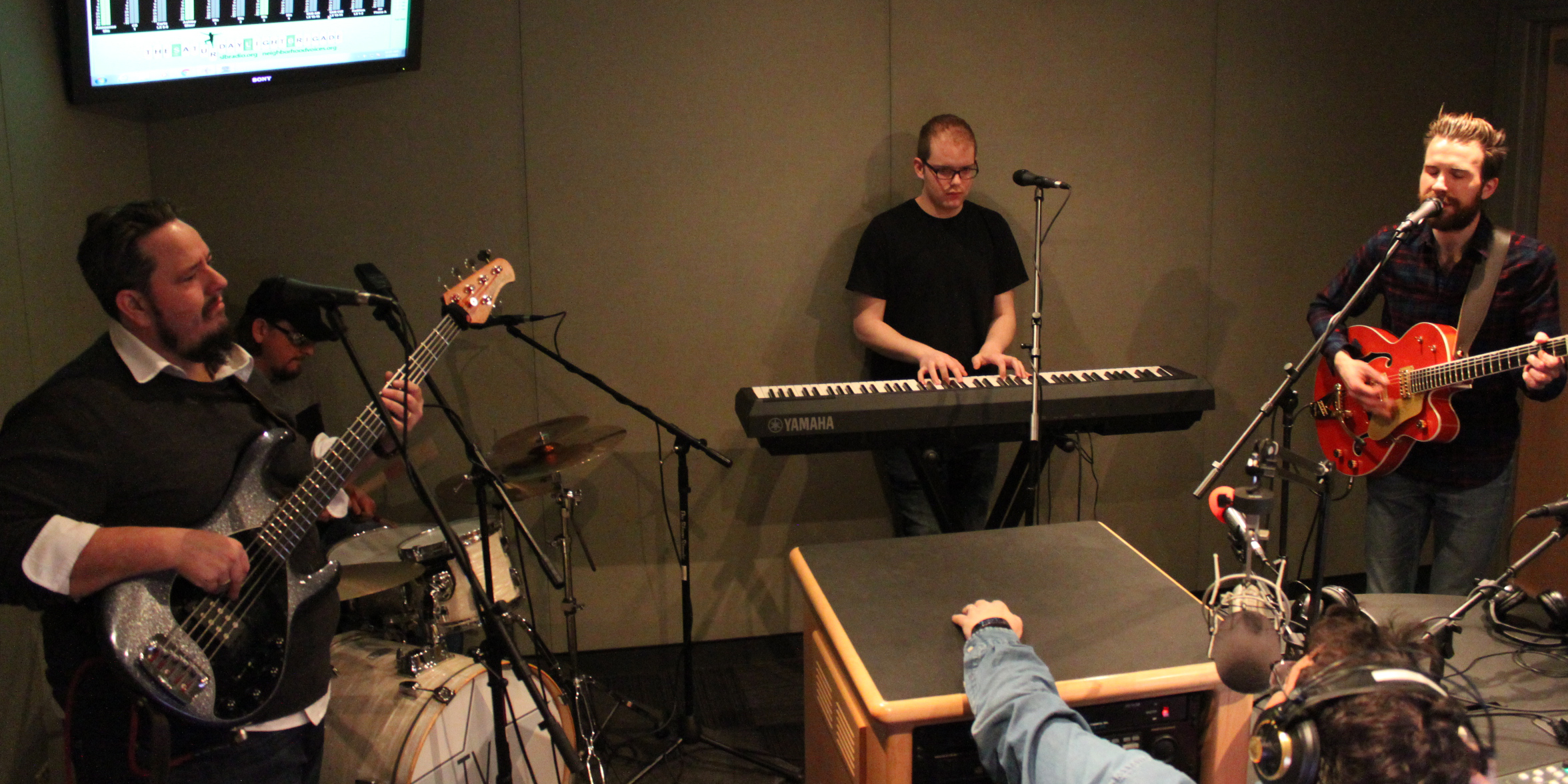 An adult playing electric bass, an adult playing a drum set, an adult playing a piano keyboard, and an adult playing electric guitar while another adult wearing headphones sitting behind a microphone listens in a radio studio