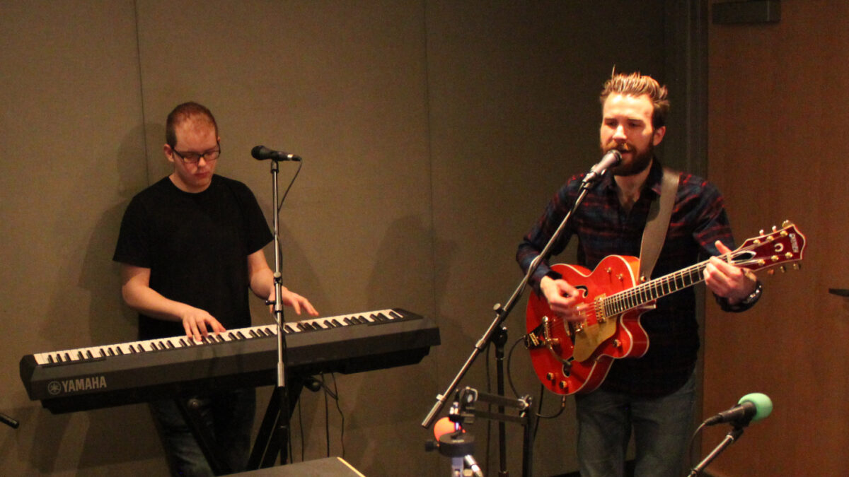 An adult playing a piano keyboard and an adult playing electric guitar and singing into a microphone in a radio studio