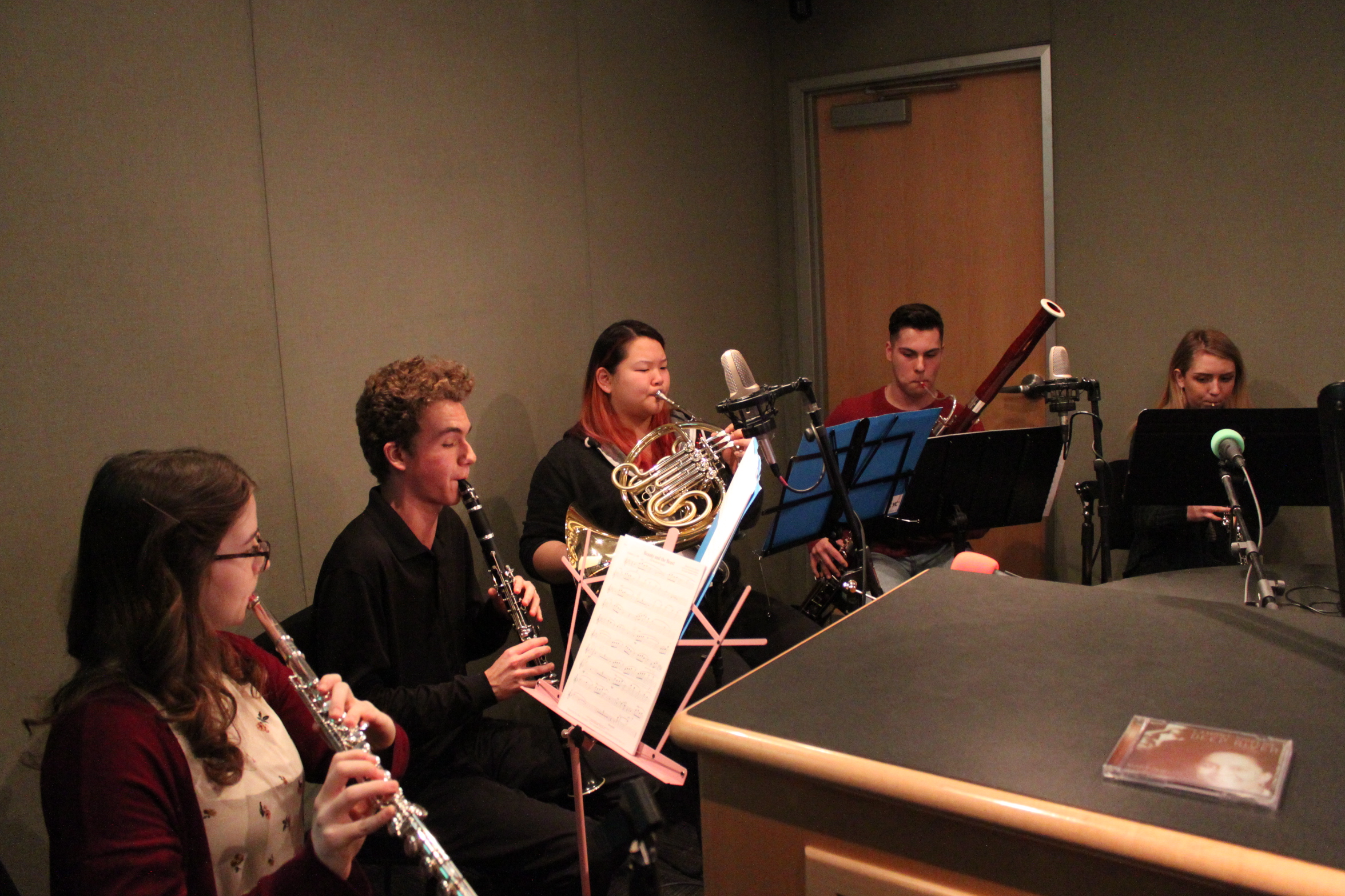 A teen playing flute, a teen playing clarinet, a teen playing French horn, a teen playing bassoon, and a teen playing oboe, all looking at sheet music on music stands in a radio studio