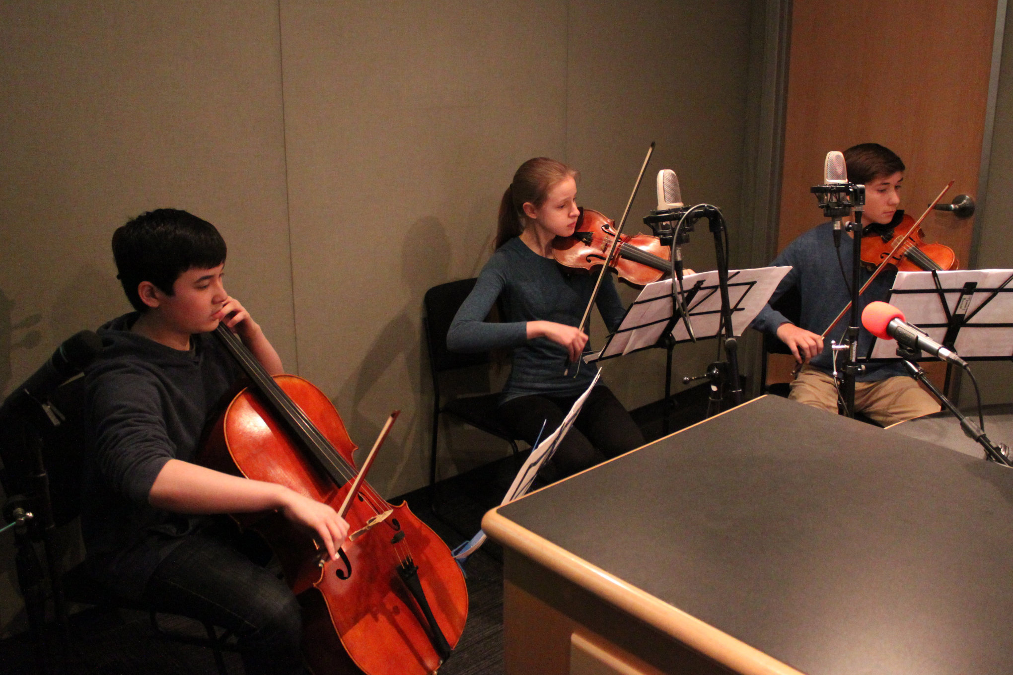 A youth playing cello and two youth playing violins, all looking at sheet music on music stands in a radio studio