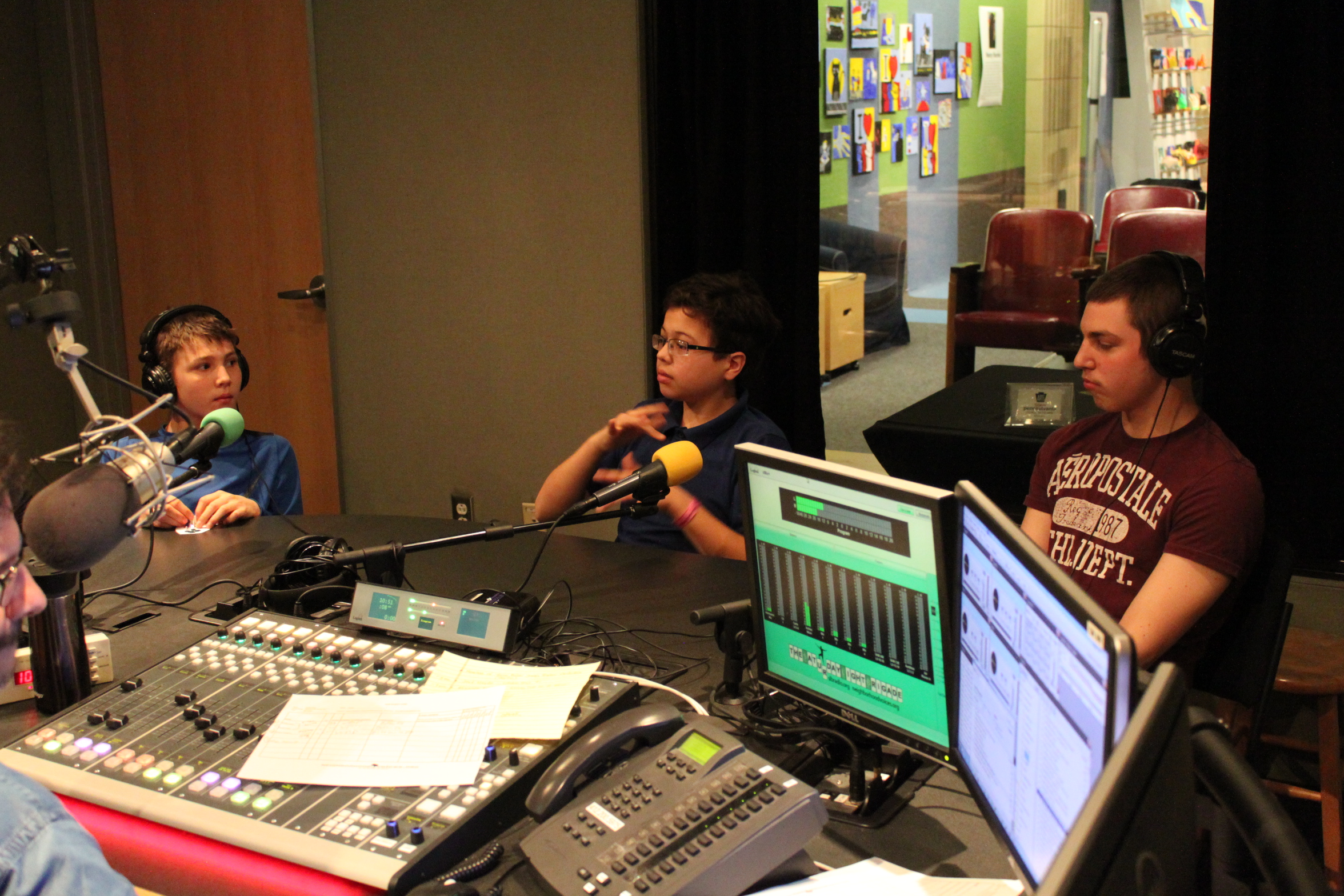 Three youth, two wearing headphones, the other gesturing with their hands, sitting behind microphones being interviewed in a radio studio