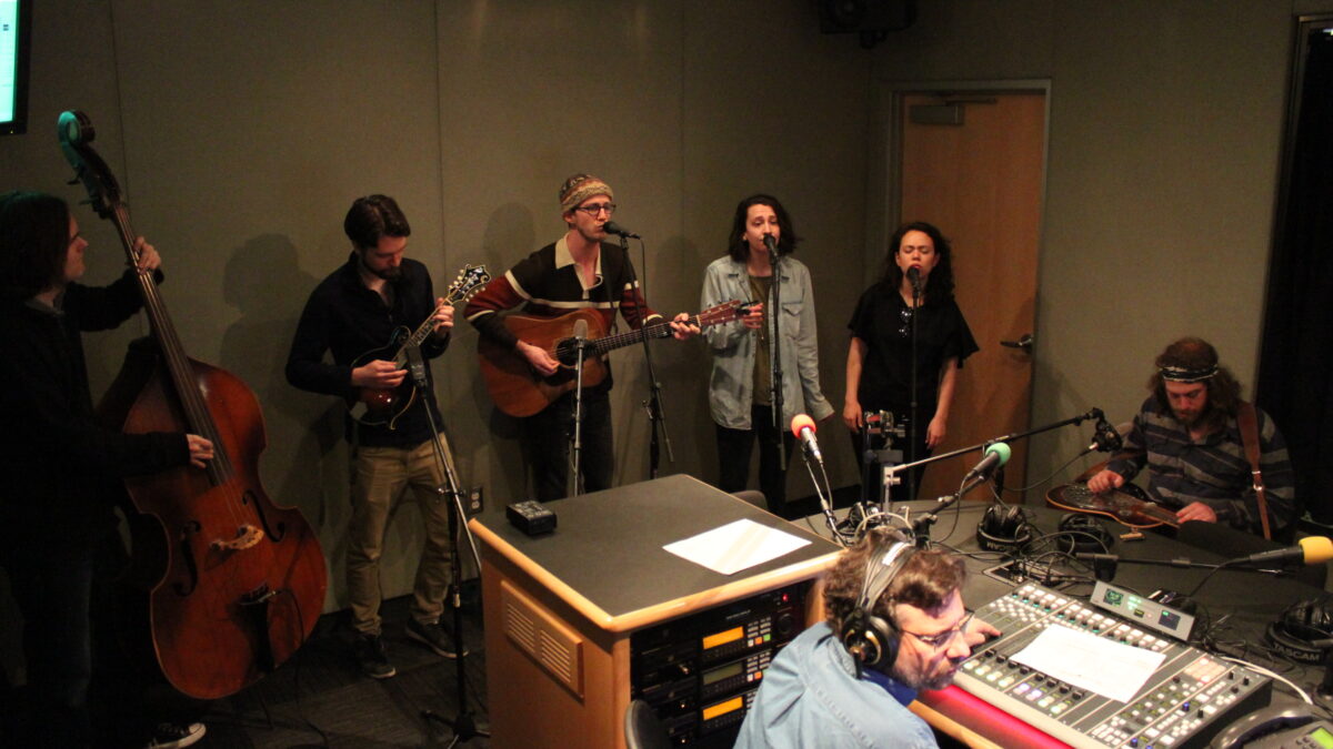 An adult playing upright bass, an adult playing mandolin, an adult playing acoustic guitar and singing into a microphone, two adults singing into microphones, and an adult playing dobro in a radio studio, as another adult wearing headphones and sitting behind a mixing board listens