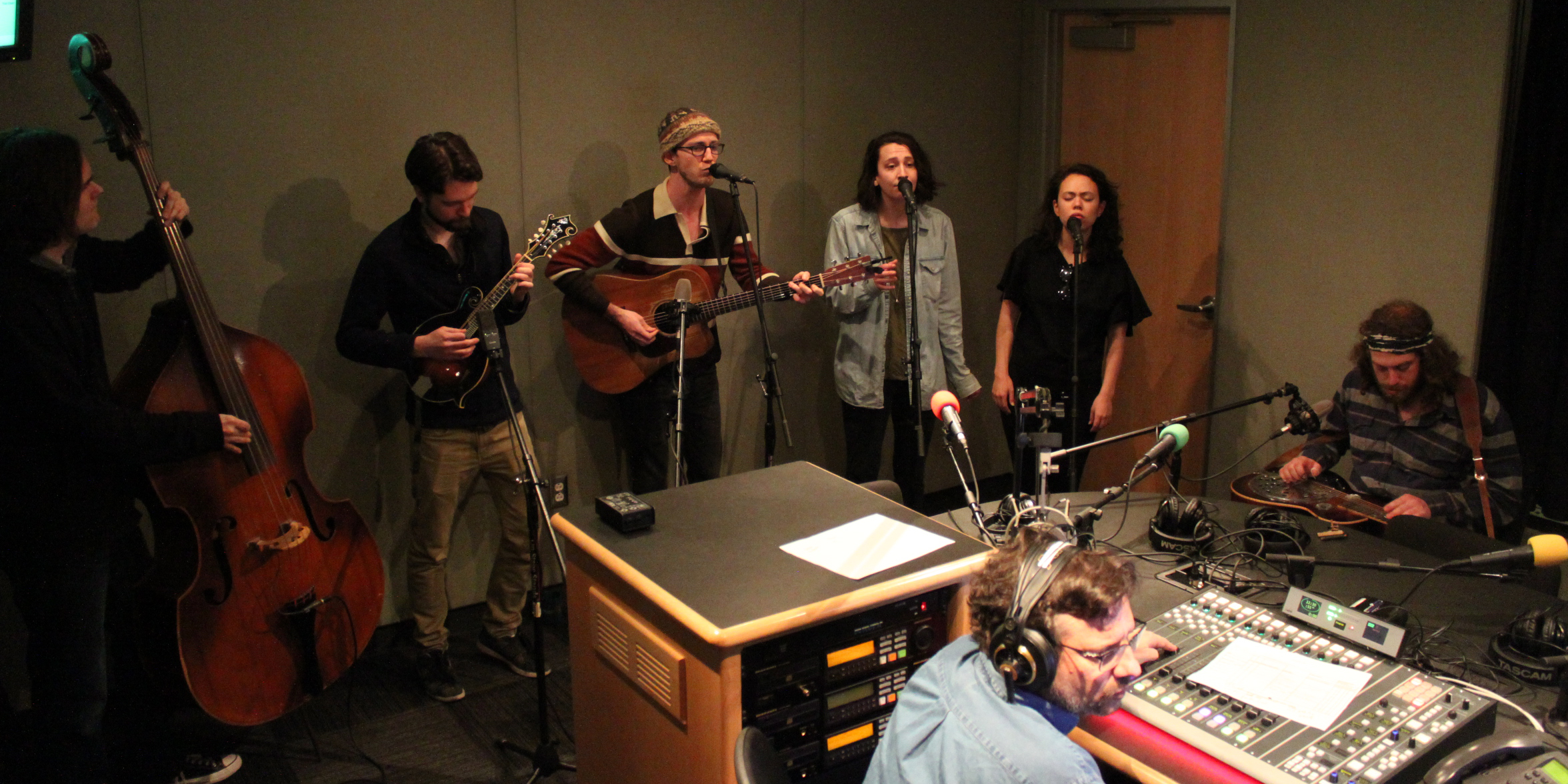 An adult playing upright bass, an adult playing mandolin, an adult playing acoustic guitar and singing into a microphone, two adults singing into microphones, and an adult playing dobro in a radio studio, as another adult wearing headphones and sitting behind a mixing board listens