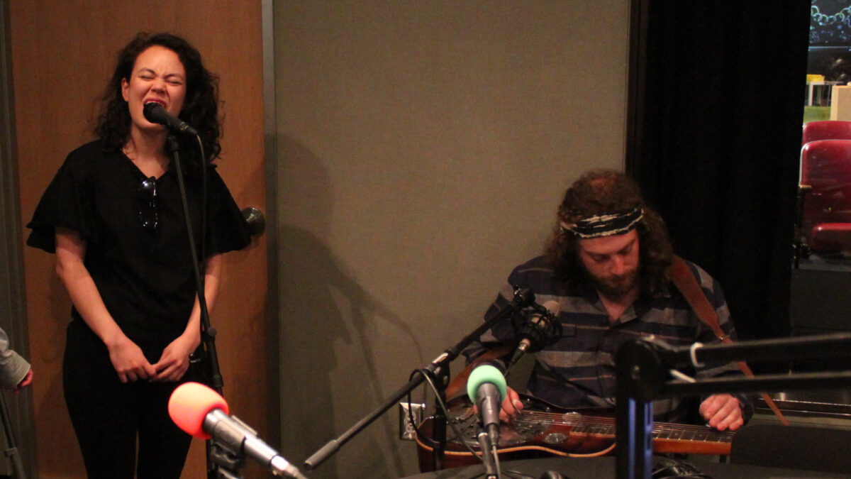 An adult singing into a microphone and an adult playing a dobro in a radio studio
