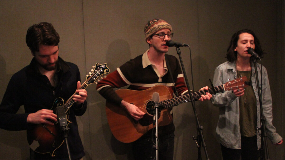 An adult playing mandolin into a microphone, an adult playing acoustic guitar and singing into a microphone, and an adult singing into a microphone in a radio studio