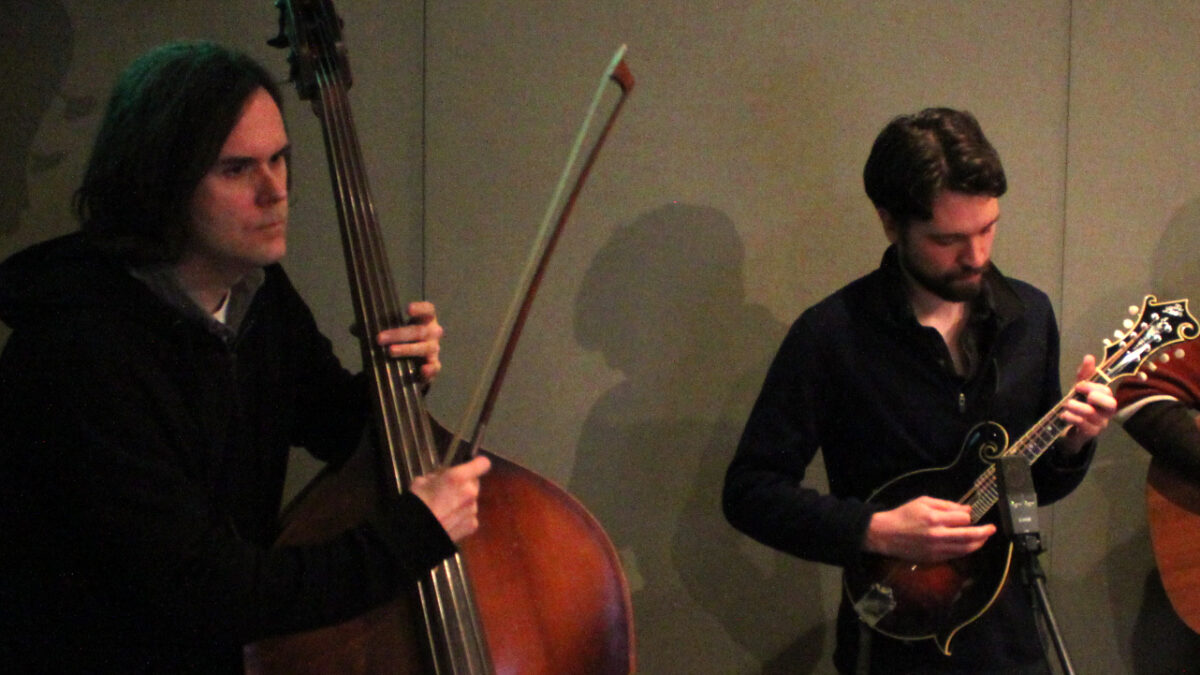 Adult playing upright bass and adult playing mandolin into a microphone in a radio studio