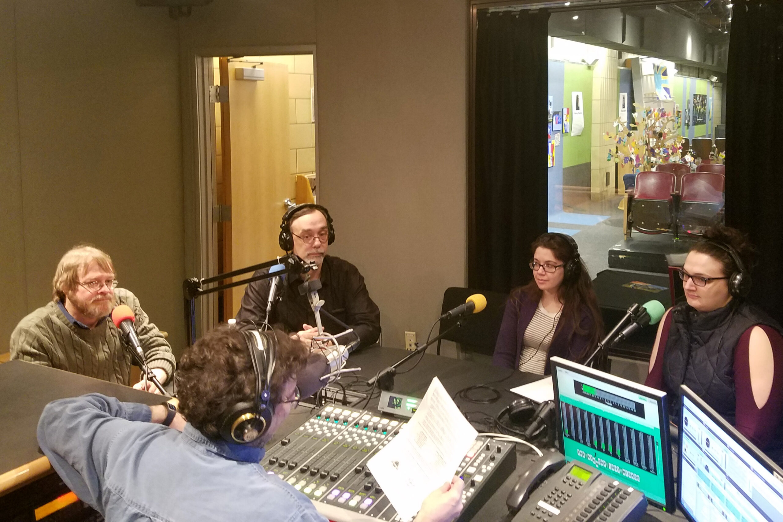 Four adults, three wearing headphones, sitting behind microphones being interviewed by an adult wearing headphones and sitting behind a microphone and a mixing board holding a piece of paper in a radio studio