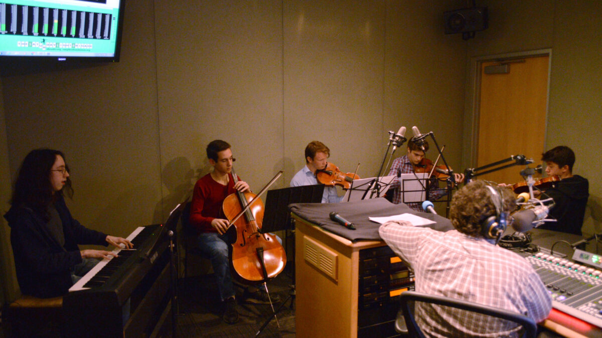 A teen playing a piano keyboard, a teen playing cello, a teen playing viola, and two teens playing violin, all looking at sheet music on music stands while an adult wearing headphones sitting behind a microphone and a mixing board listens in a radio studio