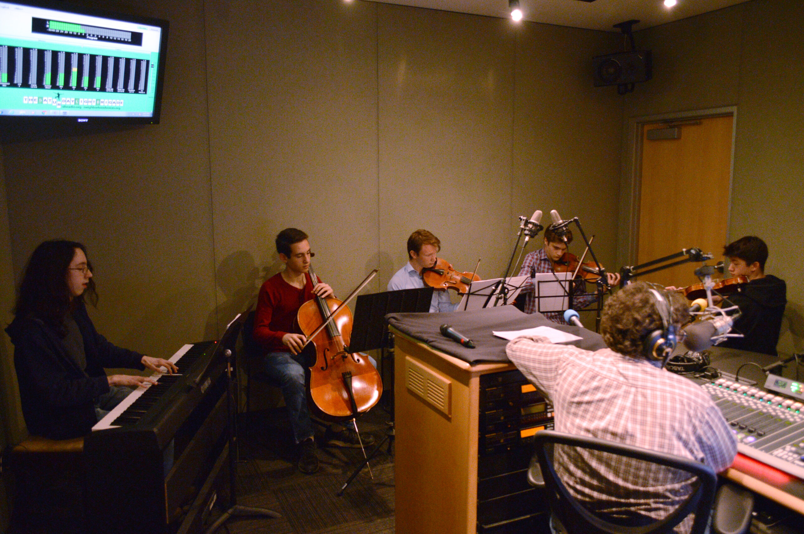 A teen playing a piano keyboard, a teen playing cello, a teen playing viola, and two teens playing violin, all looking at sheet music on music stands while an adult wearing headphones sitting behind a microphone and a mixing board listens in a radio studio