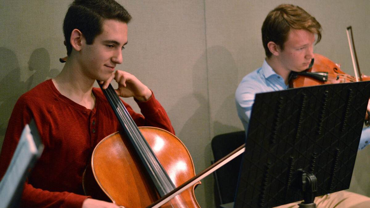 A teen playing cello and a teen playing viola, both looking at sheet music on music stands in a radio studio
