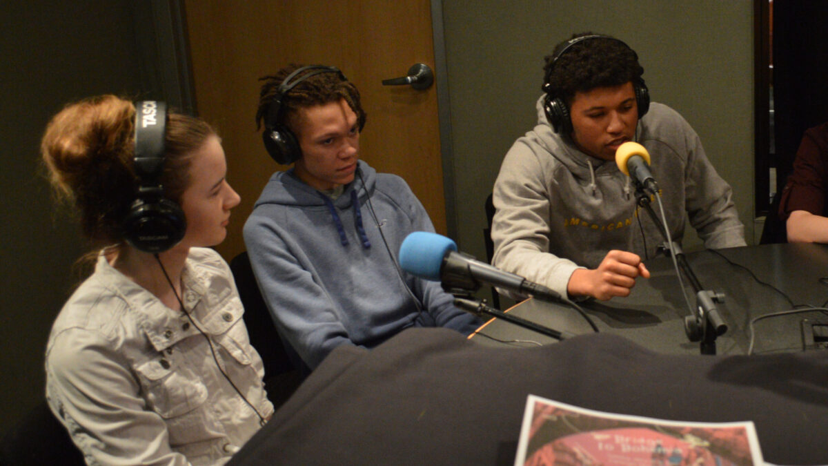 Three teens wearing headphones and speaking into microphones being interviewed in a radio studio