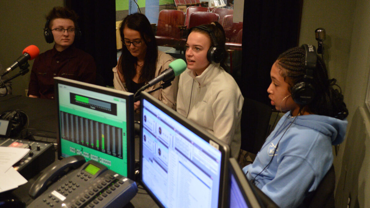 Four teens wearing headphones and speaking into microphones being interviewed in a radio studio
