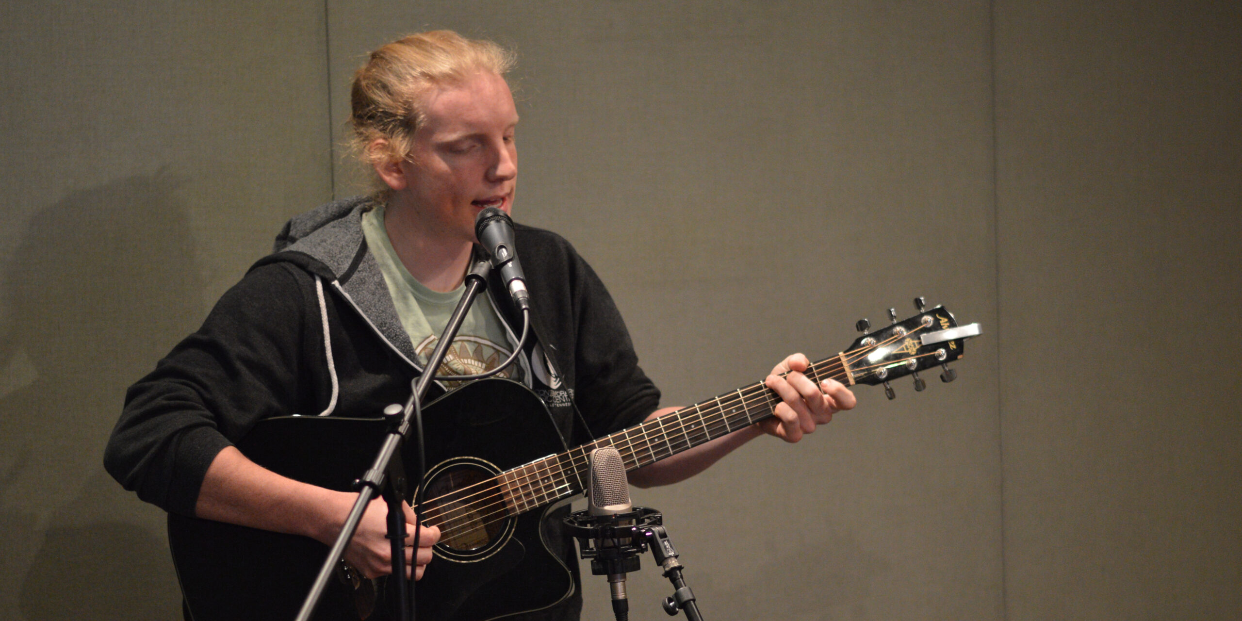 An adult playing acoustic guitar and singing into a microphone in a radio studio