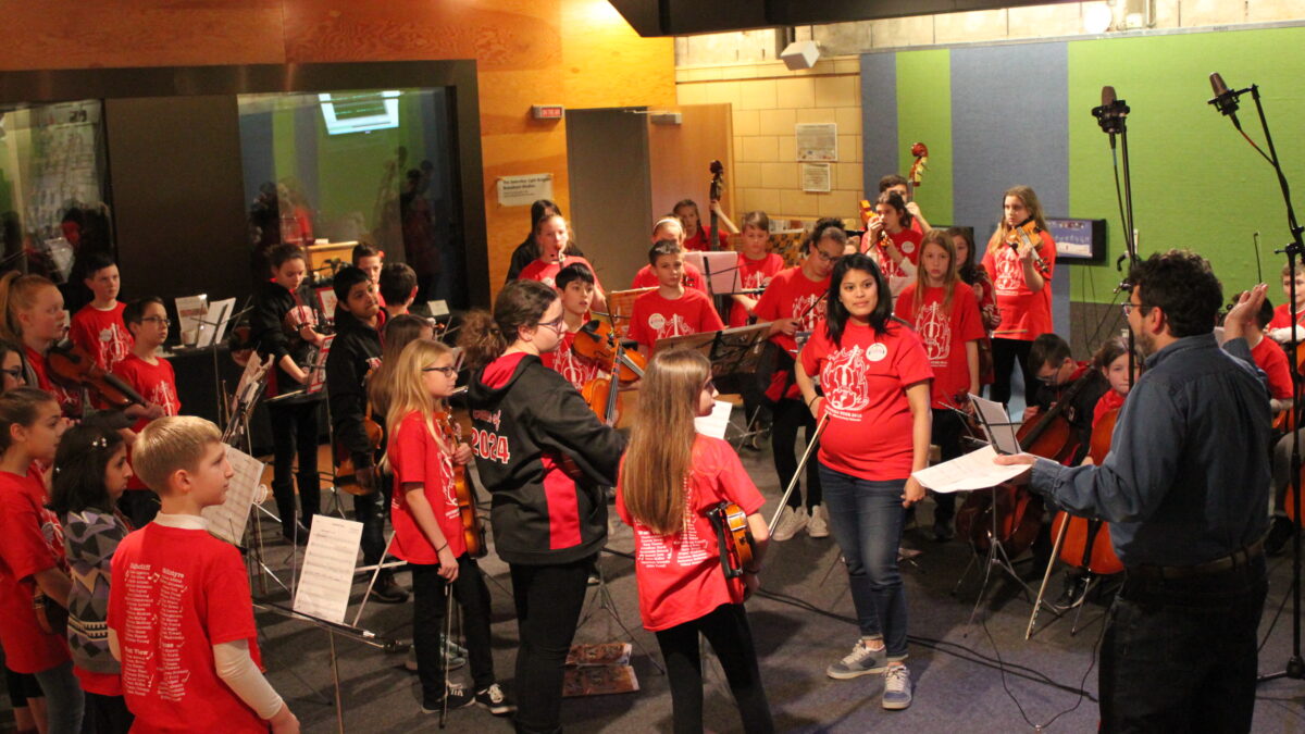 A youth orchestra holding string instruments and an adult looking at an adult gesturing with a piece of paper
