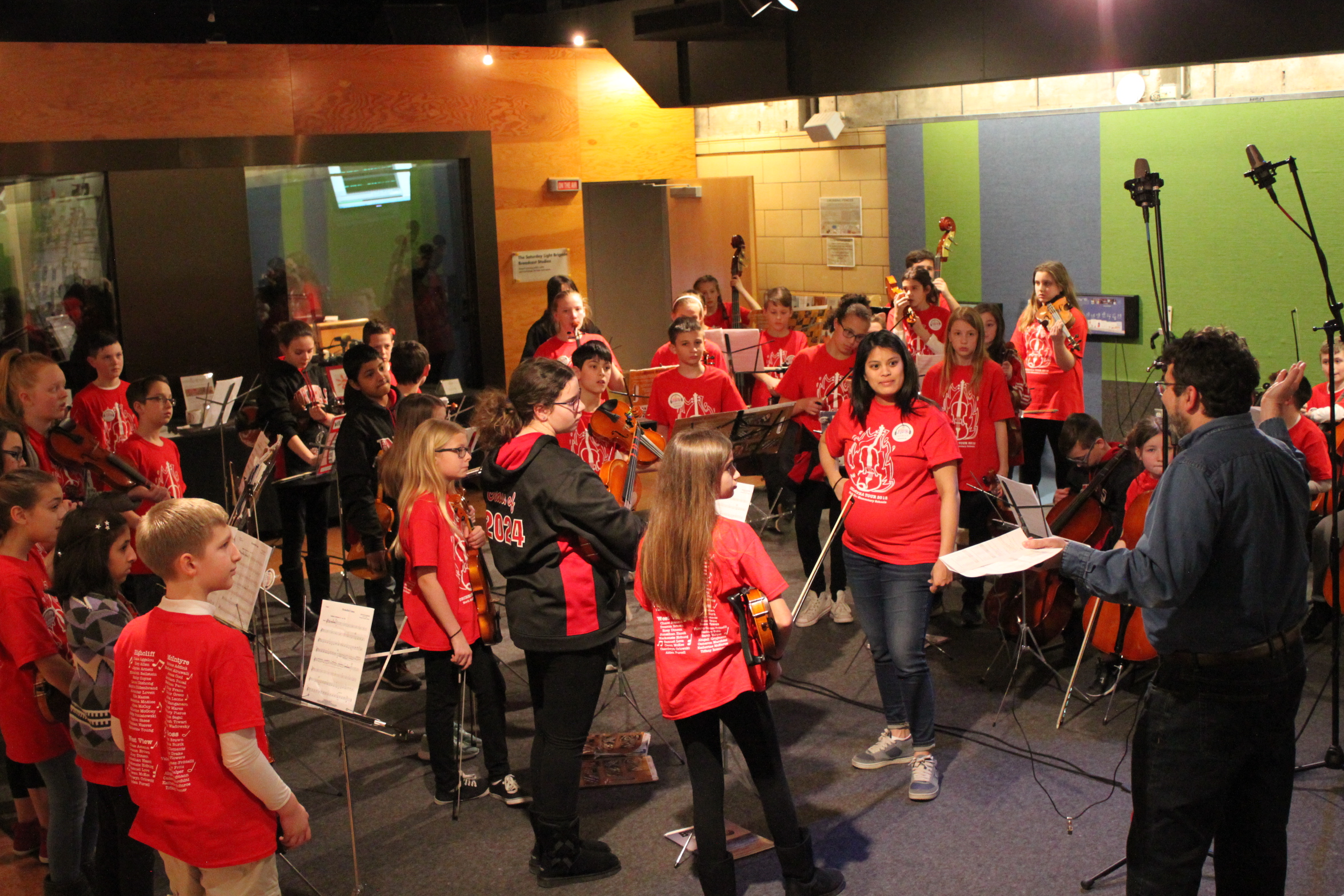 A youth orchestra holding string instruments and an adult looking at an adult gesturing with a piece of paper