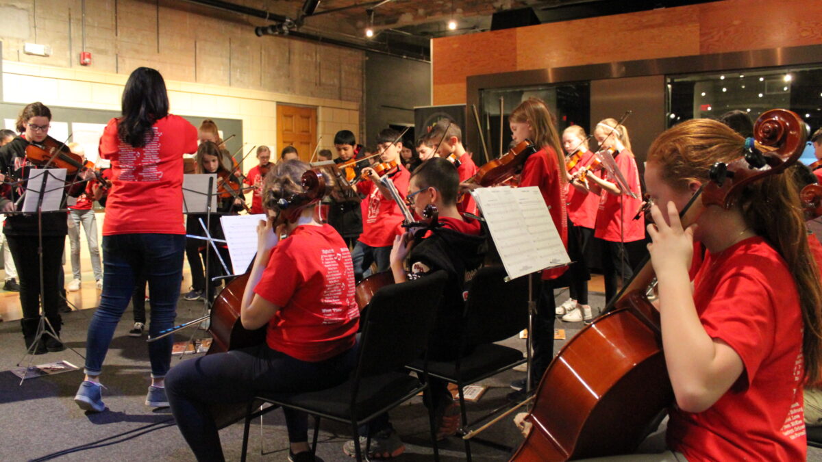 A group of youth playing string instruments in an orchestra as an adult conducts them