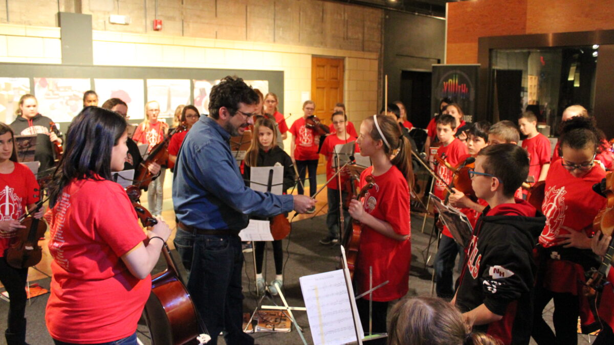 An orchestra of youth holding string instruments looking at an adult holding a long microphone interviewing a youth holding a violin
