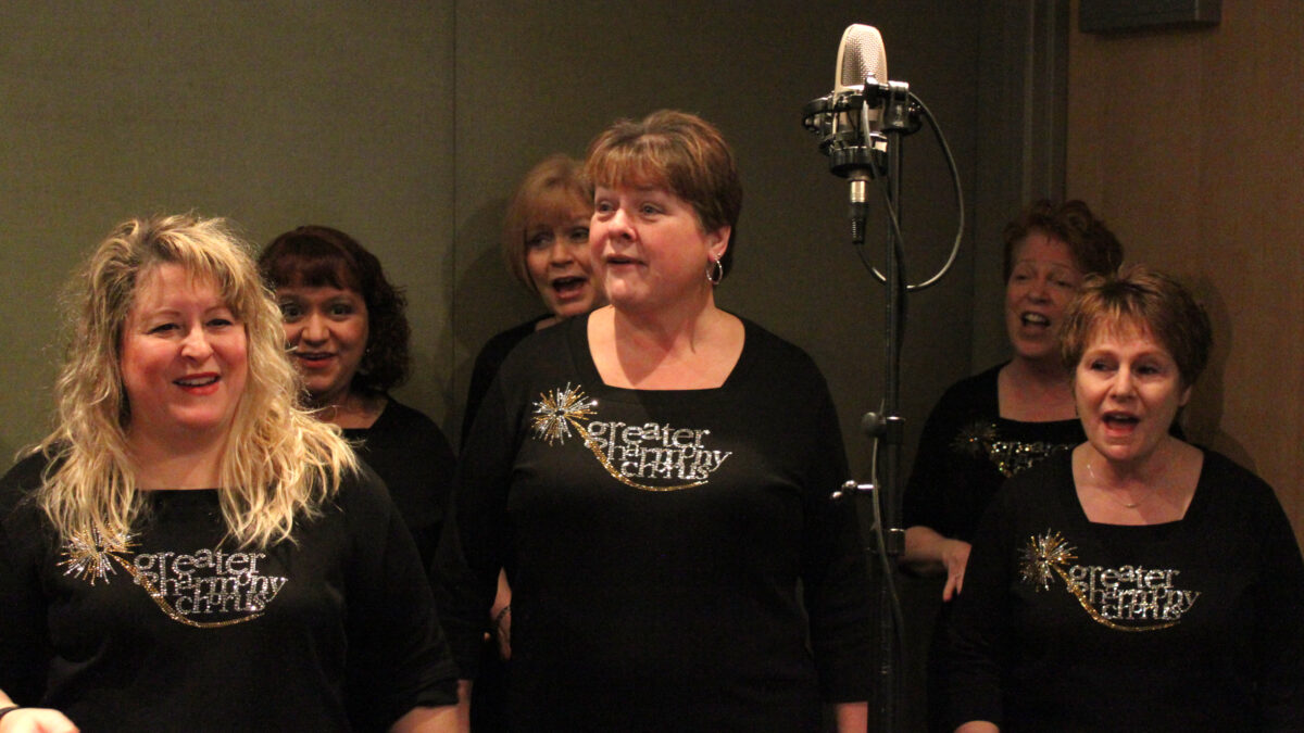 Six adults singing as a choir into a microphone in a radio studio