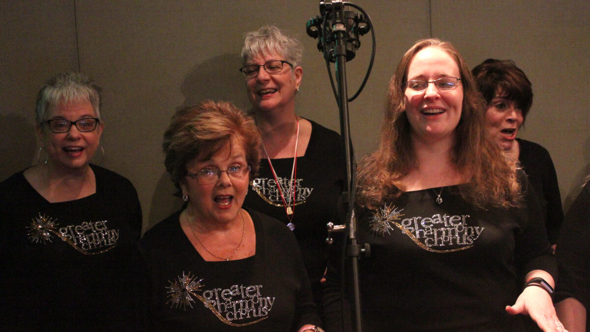 Five adults singing into a microphone as a choir in a radio studio