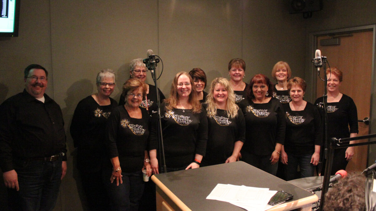 Twelve adults smiling standing behind microphones in a radio studio