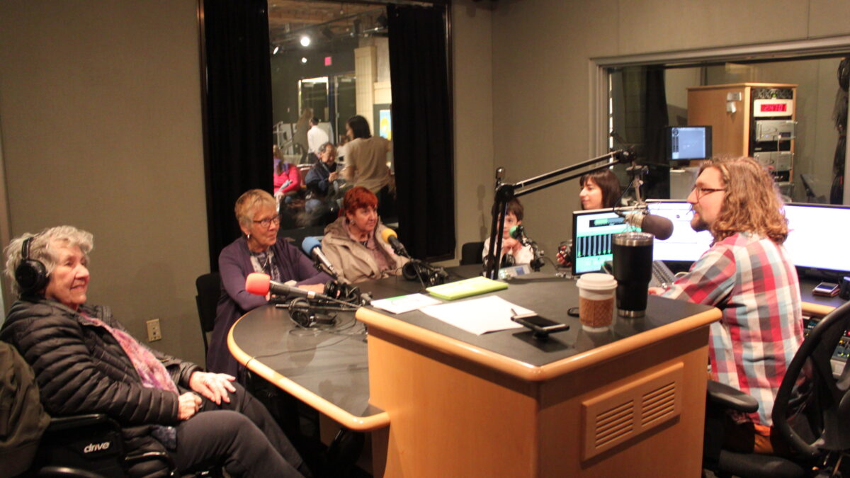 Four adults, one wearing headphones, and one child sitting behind microphones being interviewed by an adult sitting behind a microphone in a radio studio