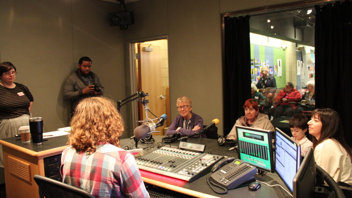 Three adults and a child sitting behind microphones being interviewed by an adult sitting behind a microphone and a mixing board while another adult prepares to take pictures and another adult supervises in a radio studio
