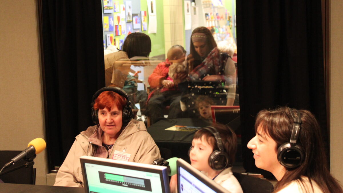 Two adults and a child wearing headphones speaking into microphones in a radio studio