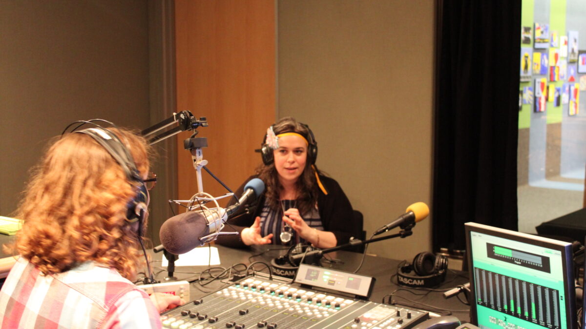 An adult wearing headphones speaking into a microphone and gesturing with their hands being interviewed by an adult wearing headphones sitting behind a microphone and a mixing board in a radio studio