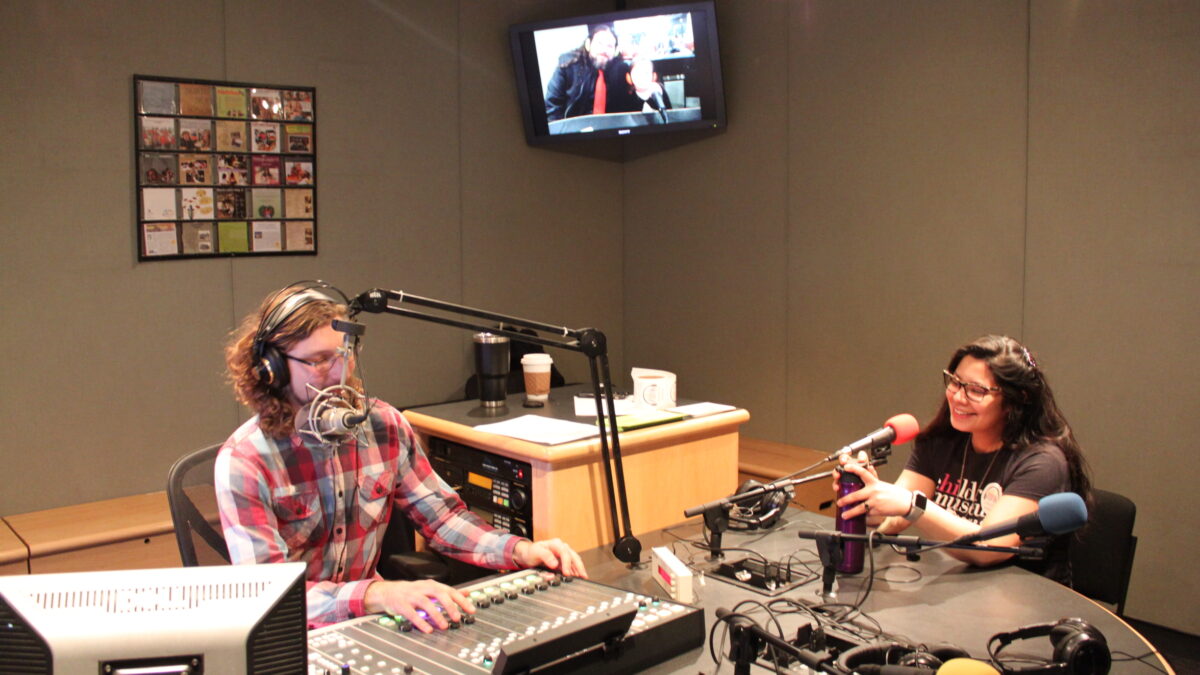 An adult smiling and sitting behind a microphone holding a water bottle being interviewed by an adult wearing headphones and sitting behind a microphone fiddling with a mixing board in a radio studio