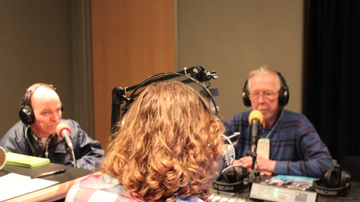 Two adults wearing headphones sitting behind microphones being interviewed by an adult wearing headphones sitting behind a microphone and a mixing board in a radio studio