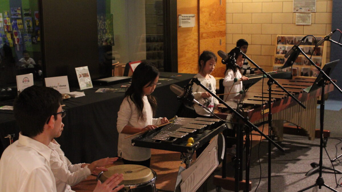 An adult playing a bongo and a youth playing a conga with drumsticks, a youth playing glockenspiel, two youth playing marimba, and a youth playing a piano keyboard