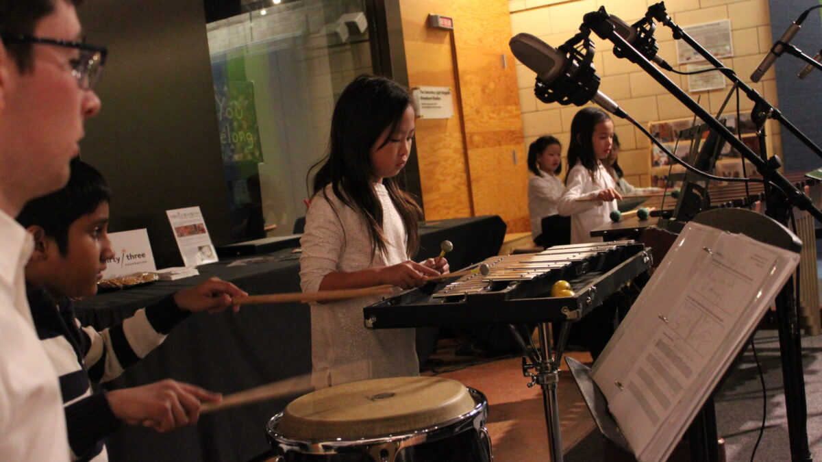 A youth playing a conga with drumsticks, a youth playing glockenspiel, a youth playing marimba, and two youth playing a piano keyboard