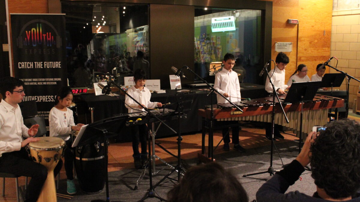 A youth playing a conga, a youth playing a bongo, a youth playing a glockenspiel, two youth playing marimba, and two youth playing a piano keyboard