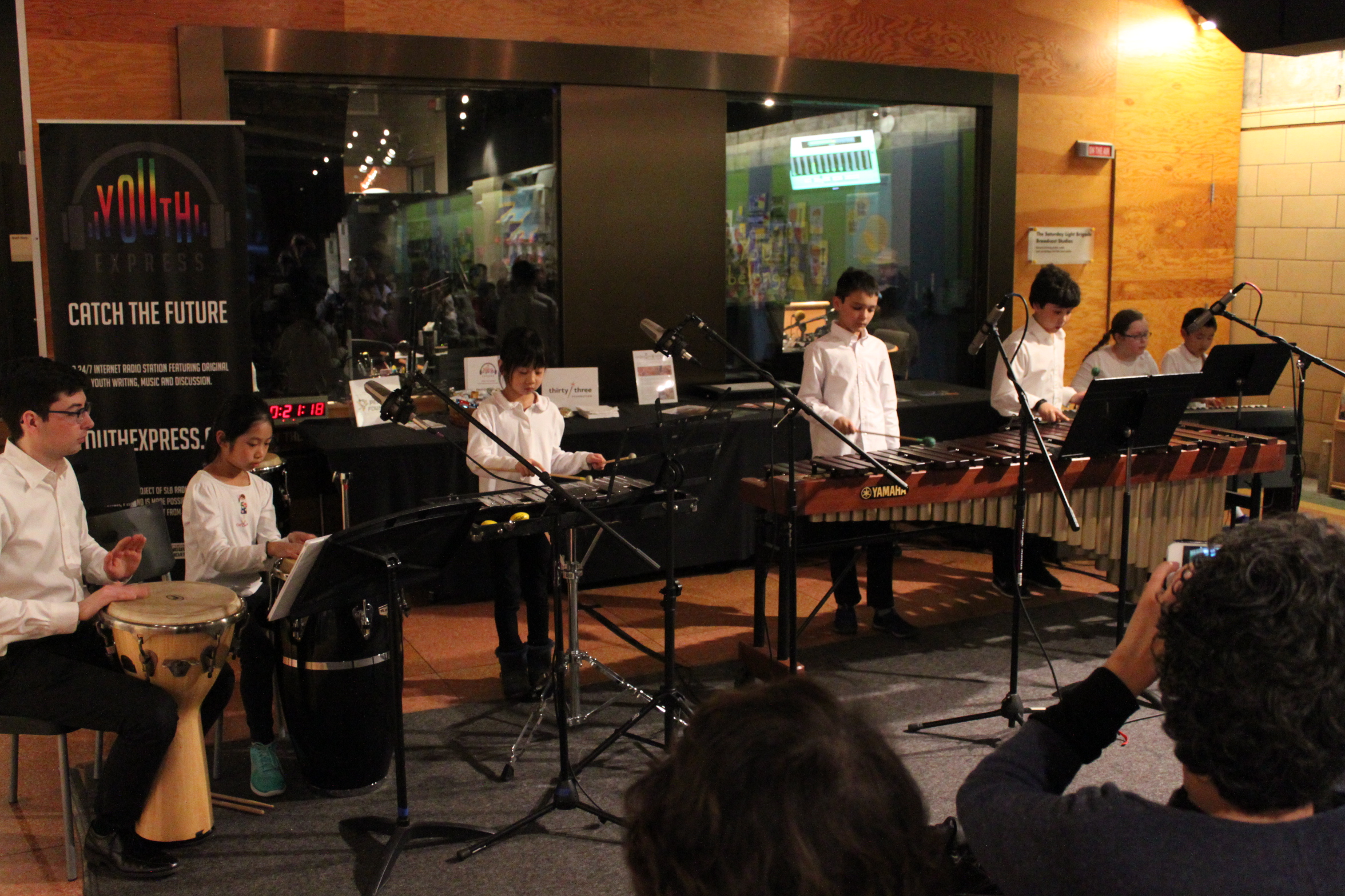 A youth playing a conga, a youth playing a bongo, a youth playing a glockenspiel, two youth playing marimba, and two youth playing a piano keyboard