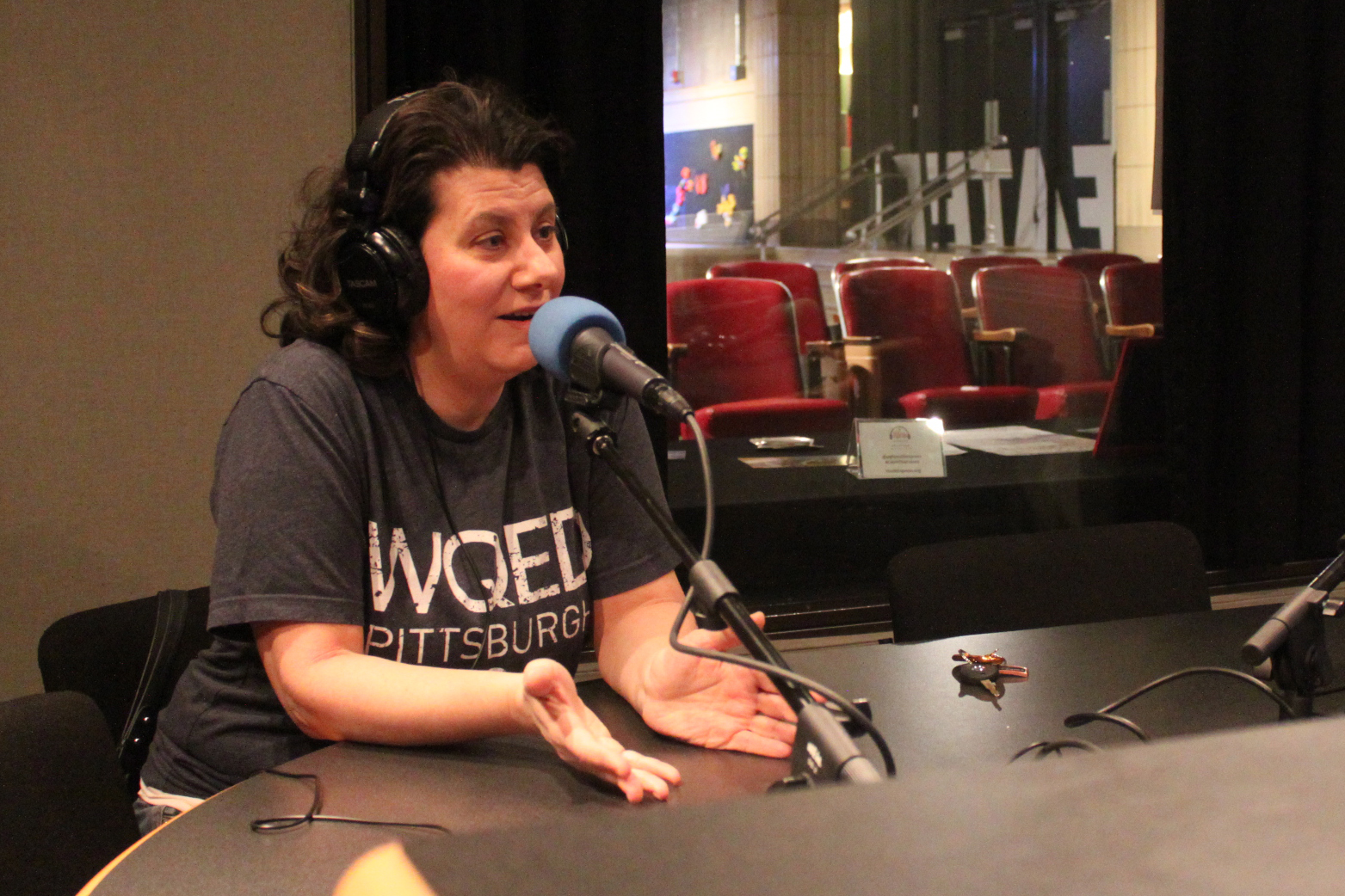 An adult wearing headphones speaking into a microphone and gesturing with their hands in a radio studio